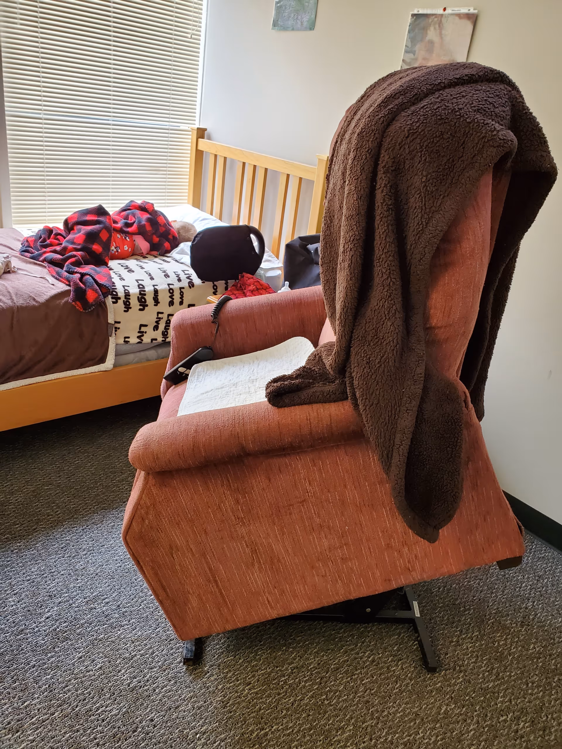 A bedroom in a senior facility with a bed, recliner, and brown blanket draped over chair.