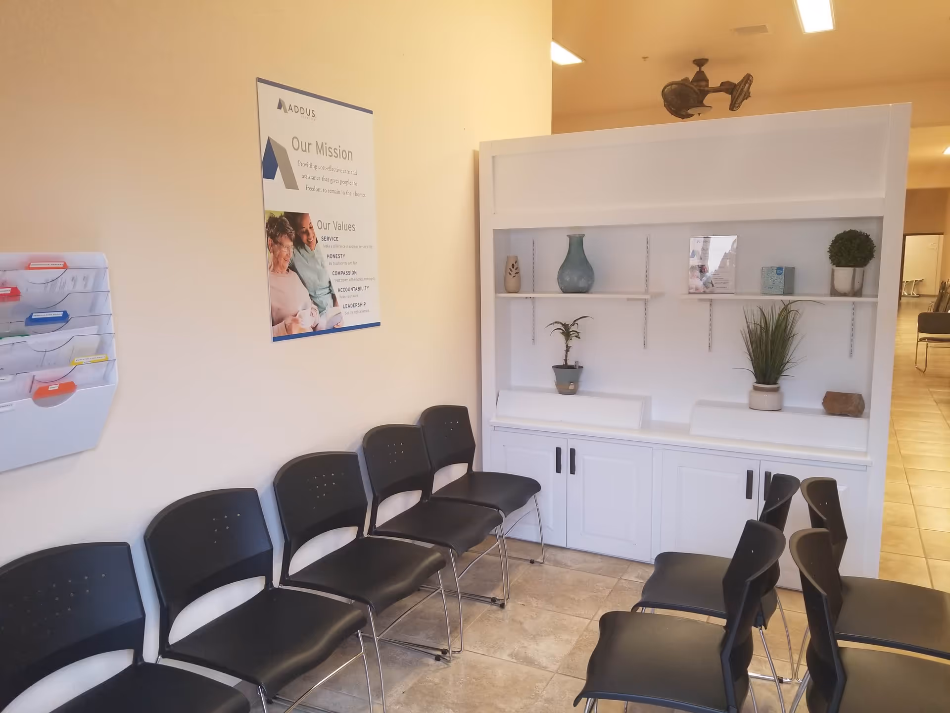 Interior waiting area with black chairs, a white shelving unit, and a wall poster.