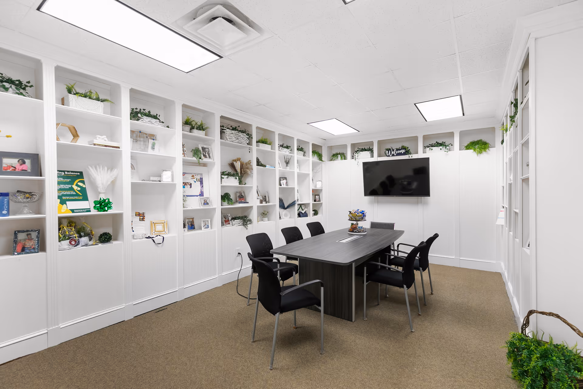 Bright white meeting room with built-in shelves, a central table, and a wall-mounted TV.