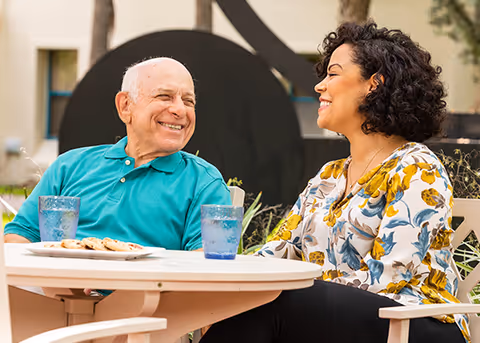 Two people sit at an outdoor table with cookies and drinks, smiling.