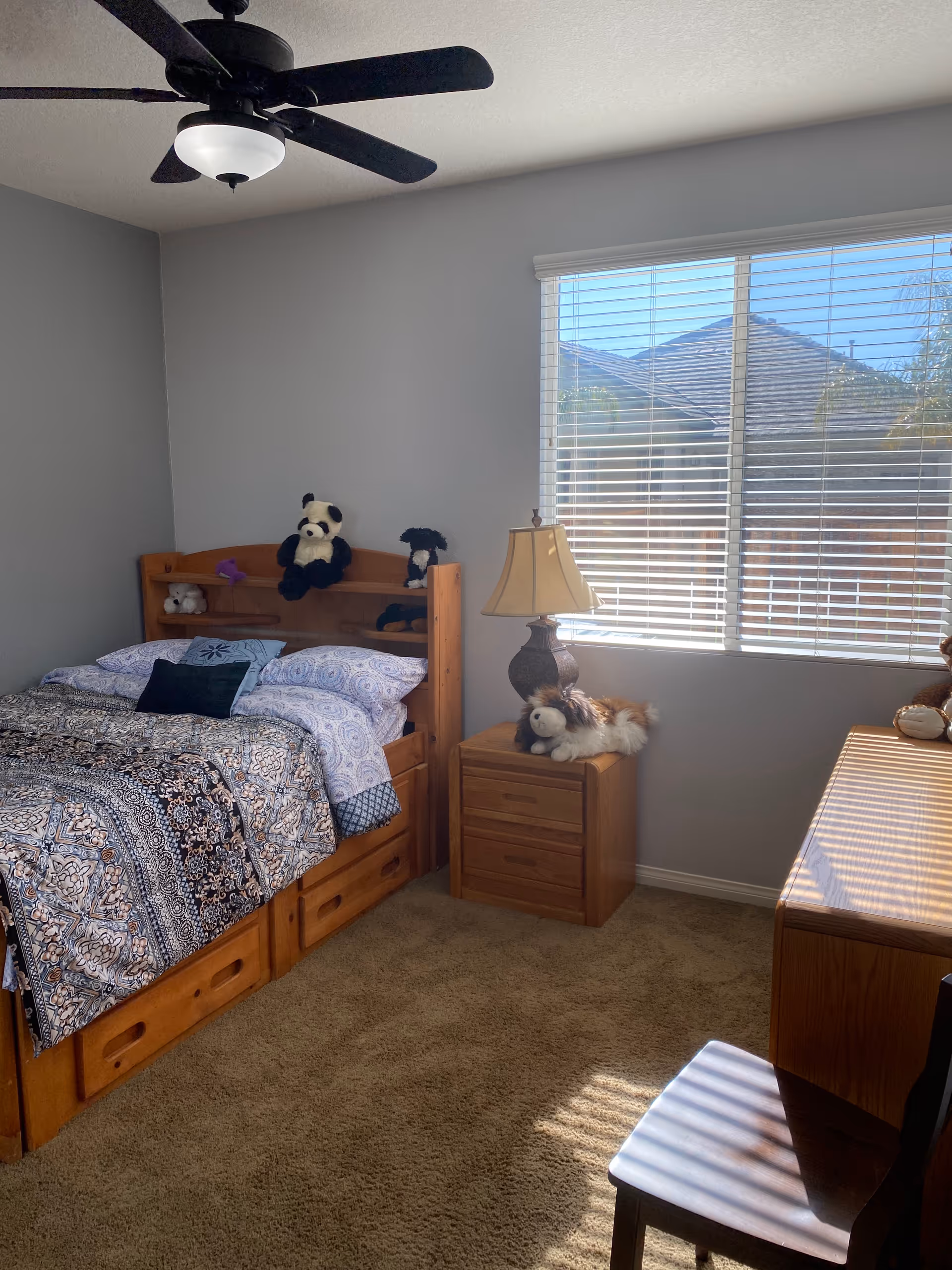 Wooden twin bed with patterned bedding, nightstand, lamp, and window blinds.
