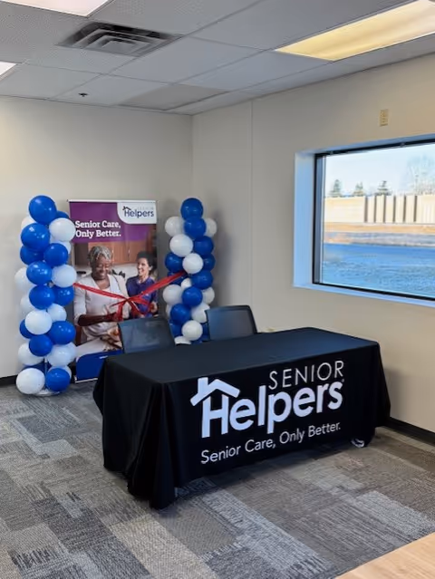 Indoor event setup with a Senior Helpers banner, balloon columns, and a branded table.