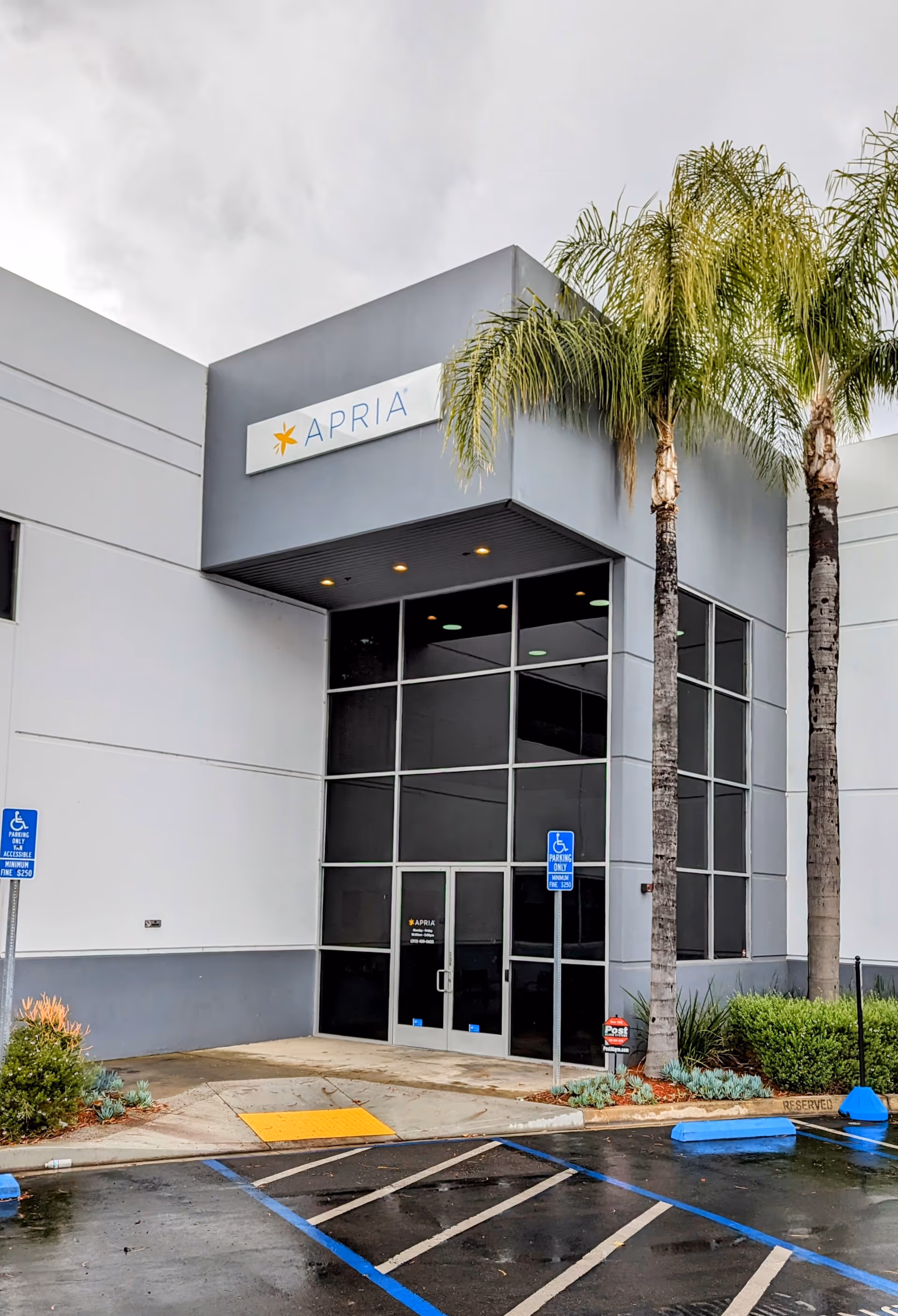 Modern healthcare facility entrance with glass doors, APRIA sign, and palm trees.