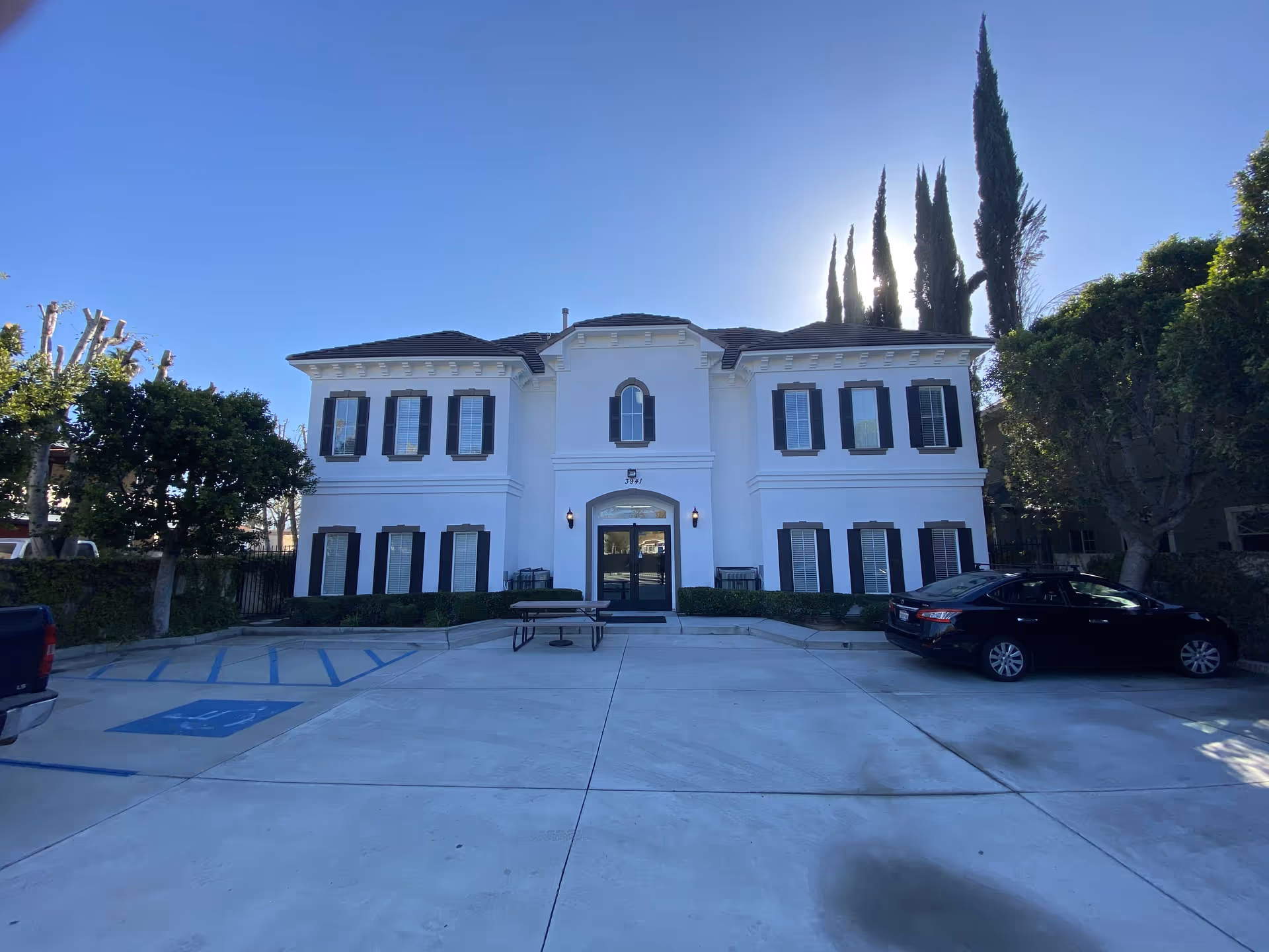 Front view of a two-story white senior living building with dark shutters and parked cars.