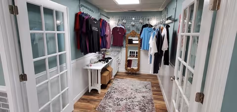 Narrow interior closet/boutique area with hanging clothes, racks, and a mirror.