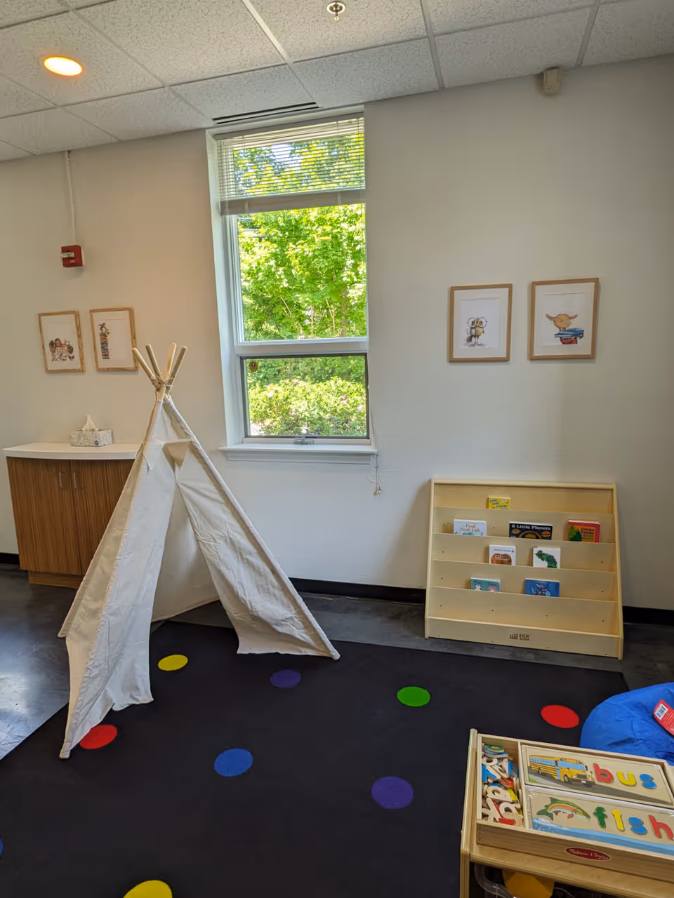 Bright indoor play area with a white teepee, bookshelf, framed art, and colorful dot carpet.