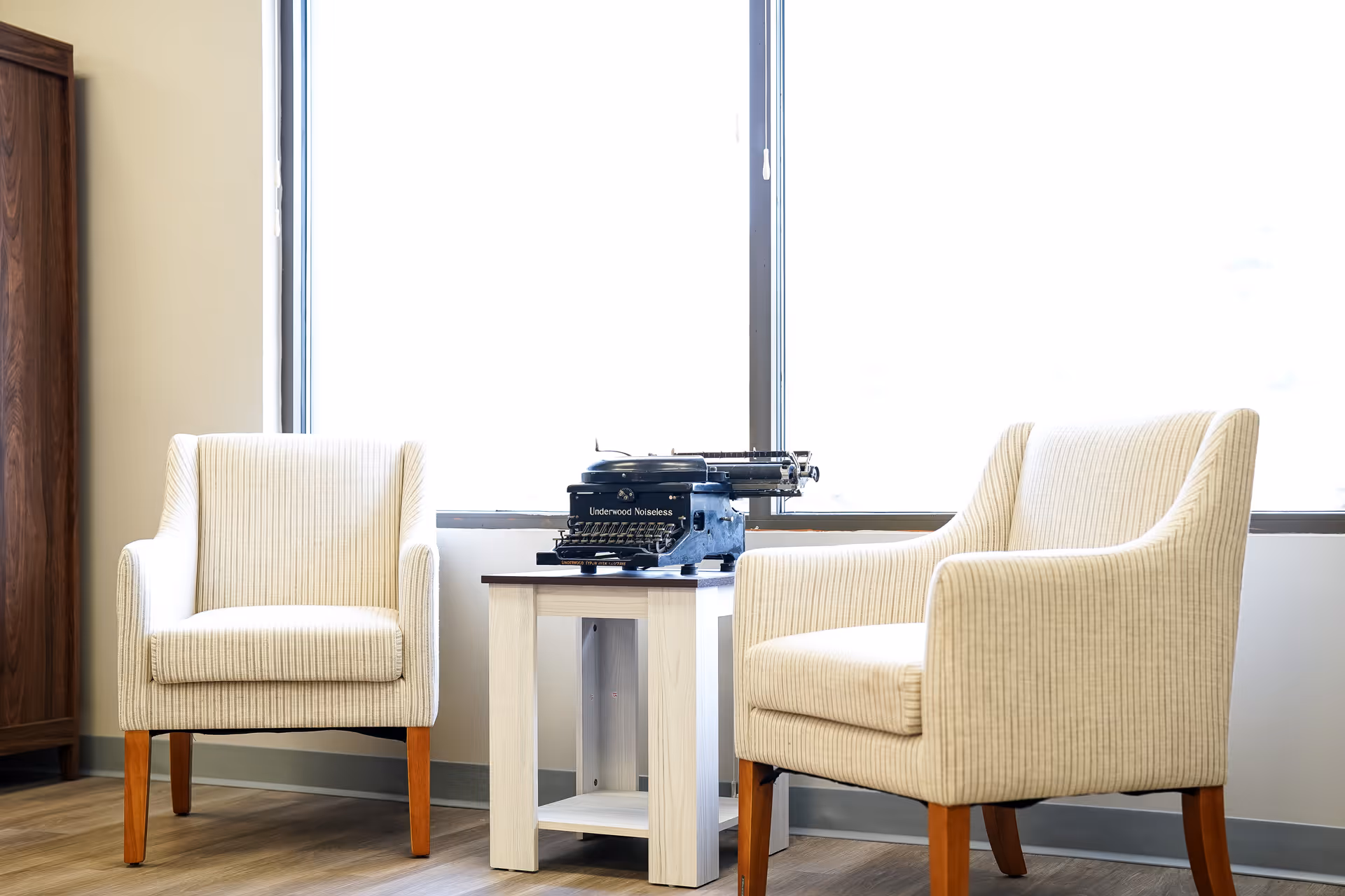 Two cream chairs flank a small table with a vintage typewriter by a bright window.