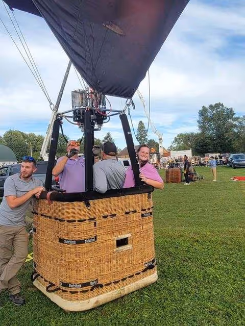 People stand around a hot air balloon basket on a grassy outdoor field.