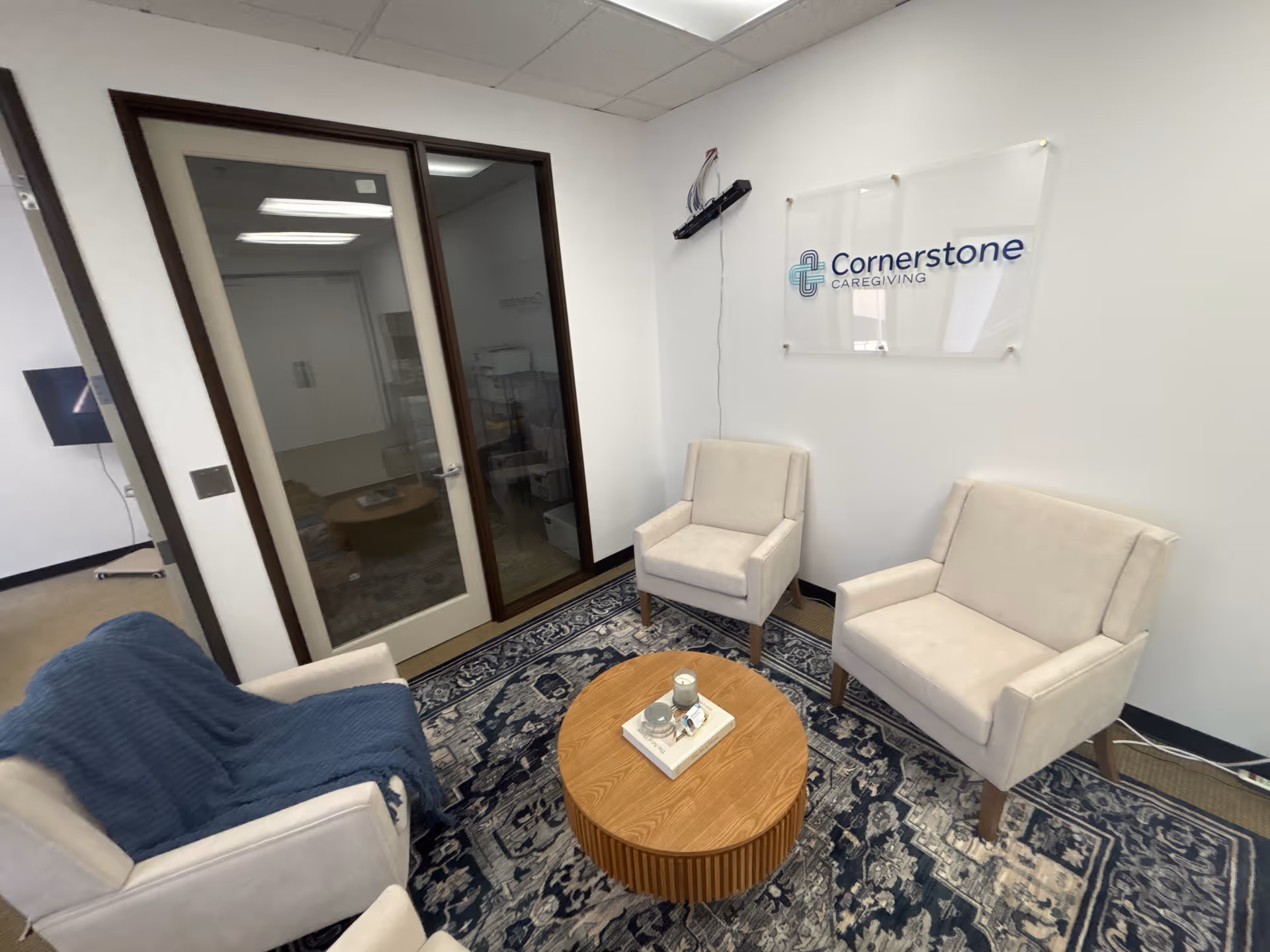 Interior waiting area with white chairs, a rug, round coffee table, and a Cornerstone Caregiving sign.