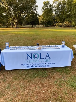 Outdoor table with NOLA @ Home Care banner on a grassy field.