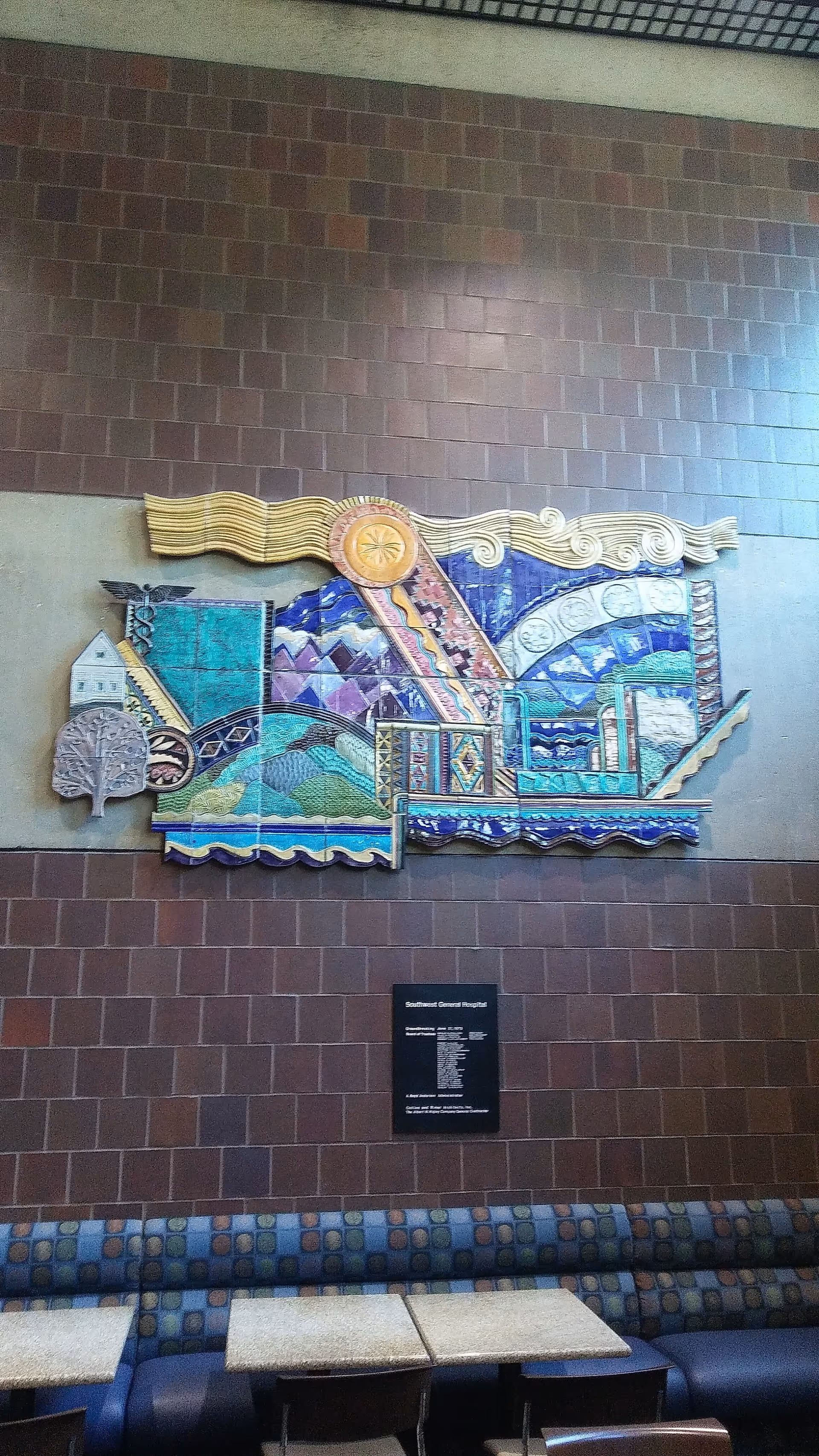 Colorful mosaic mural mounted on brown tiled interior wall above blue patterned seating.