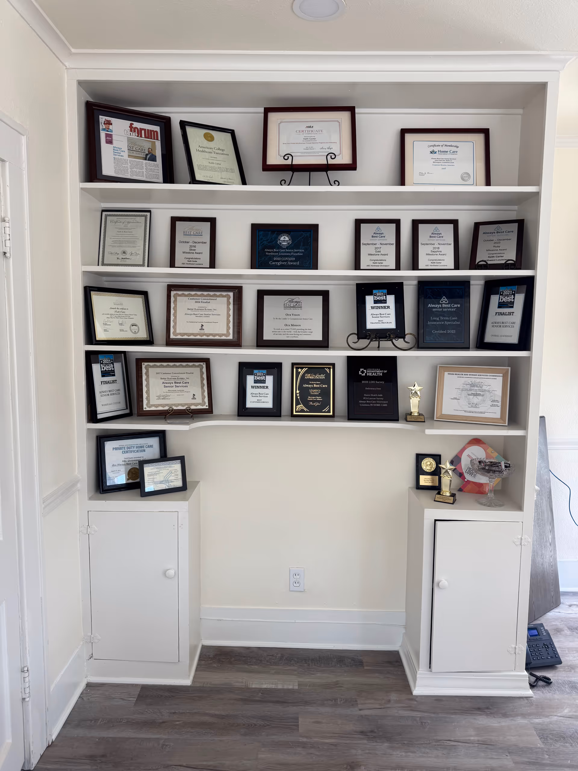 White built-in shelves displaying numerous certificates and awards in a tidy office alcove.
