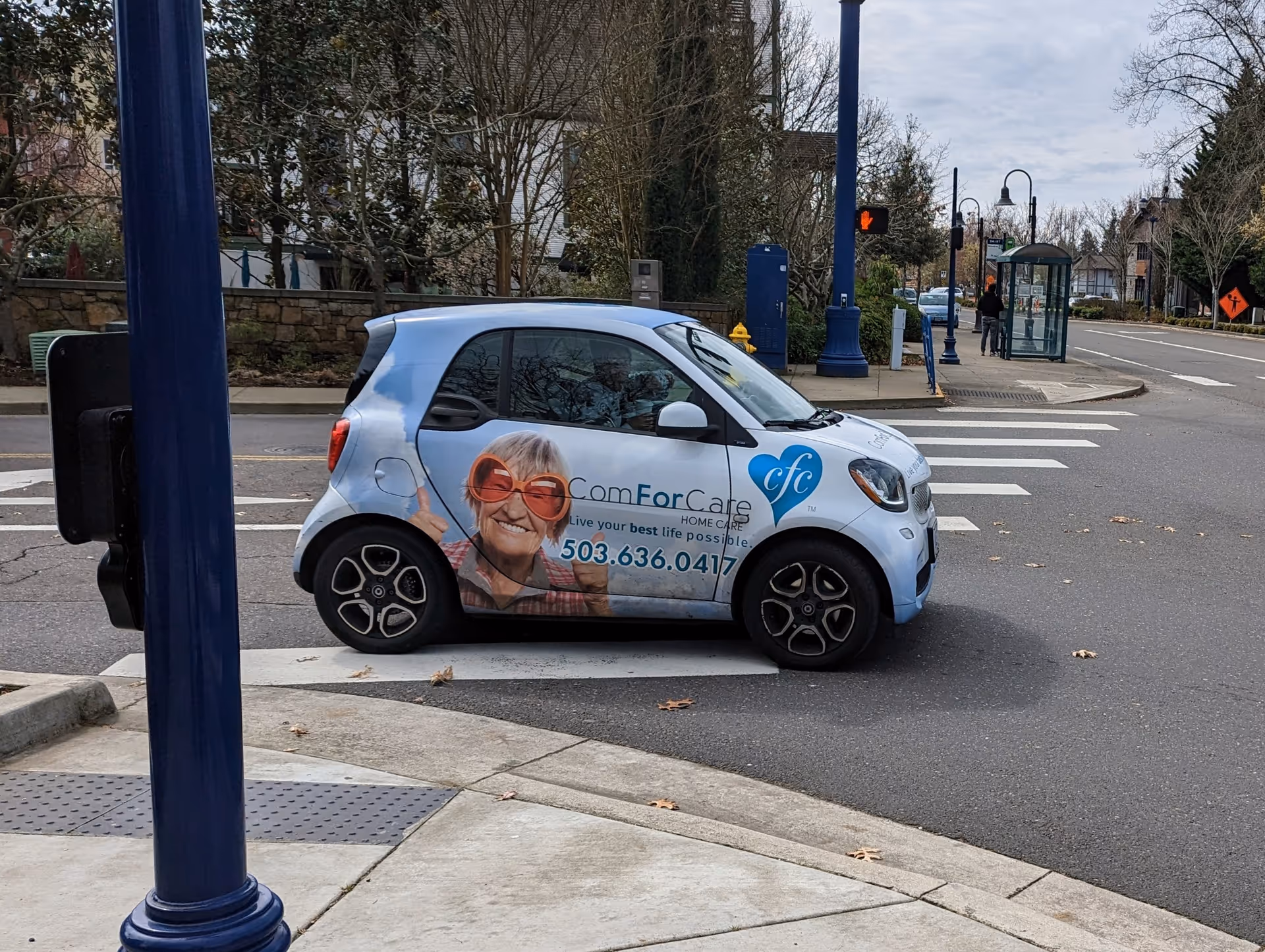 A blue ComForCare West Linn service car with an elderly person photo parked on a street.