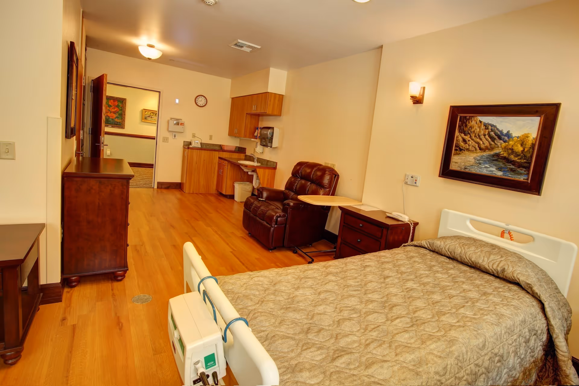 Hospice bedroom with a hospital bed, recliner, dresser, and kitchenette in warm wood tones