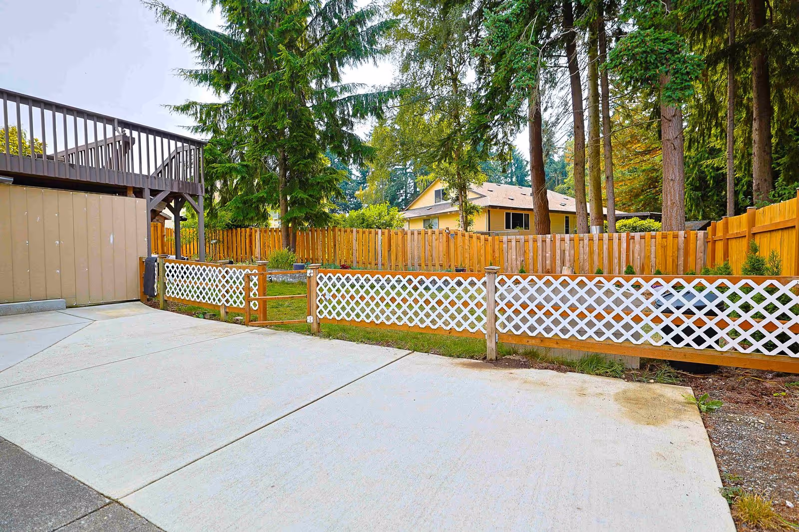 Backyard patio area with wooden lattice fence and tall trees surrounding a beige building.