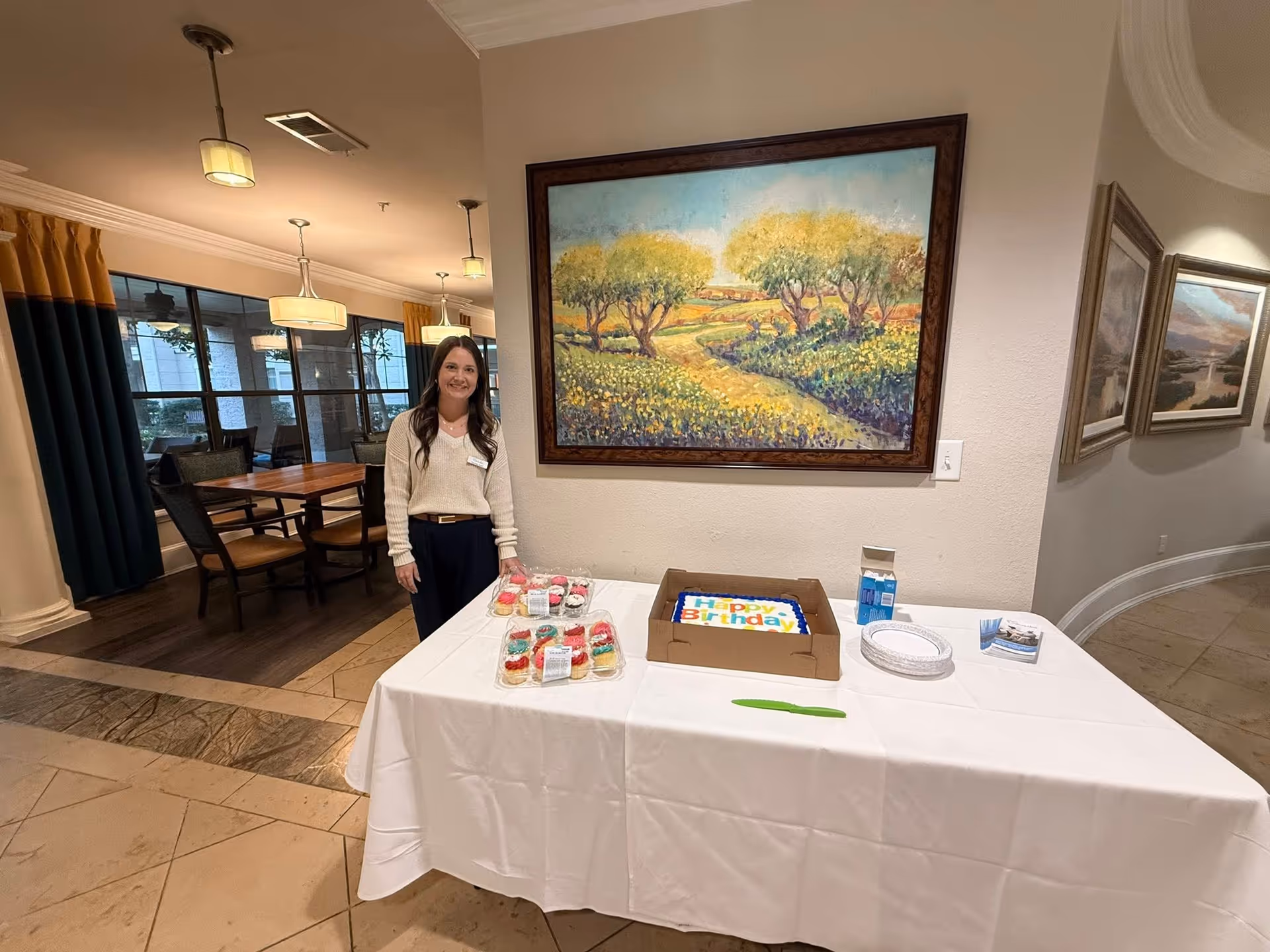 A woman stands beside a birthday cake on a table in a bright indoor lounge area.