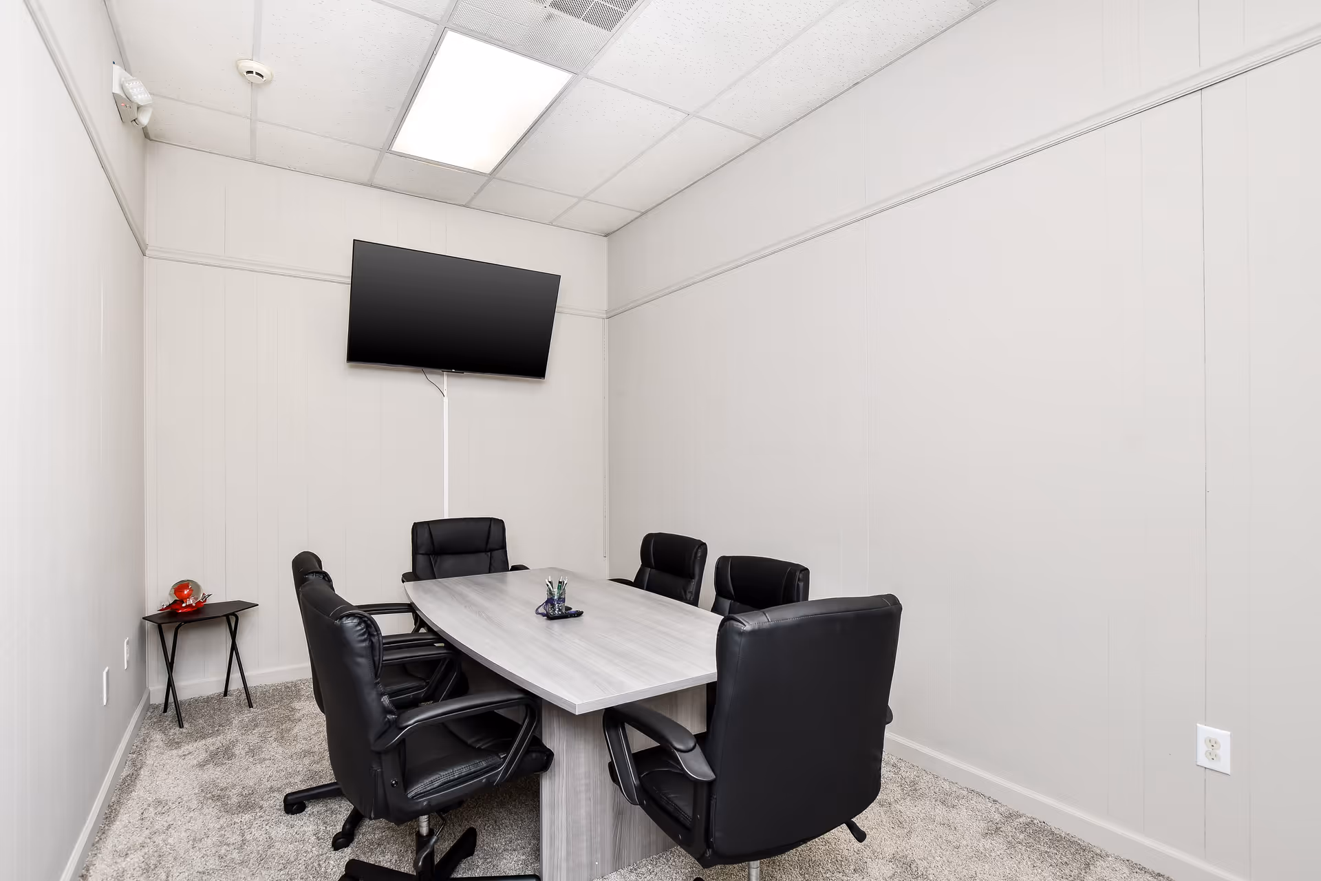 A small conference room with an oval wooden table, black chairs, and a wall-mounted TV.