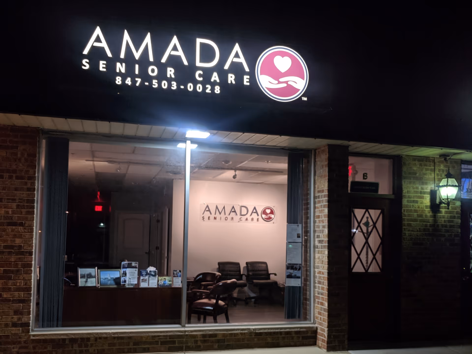 Nighttime storefront of Amada Senior Care with seating visible inside through window.