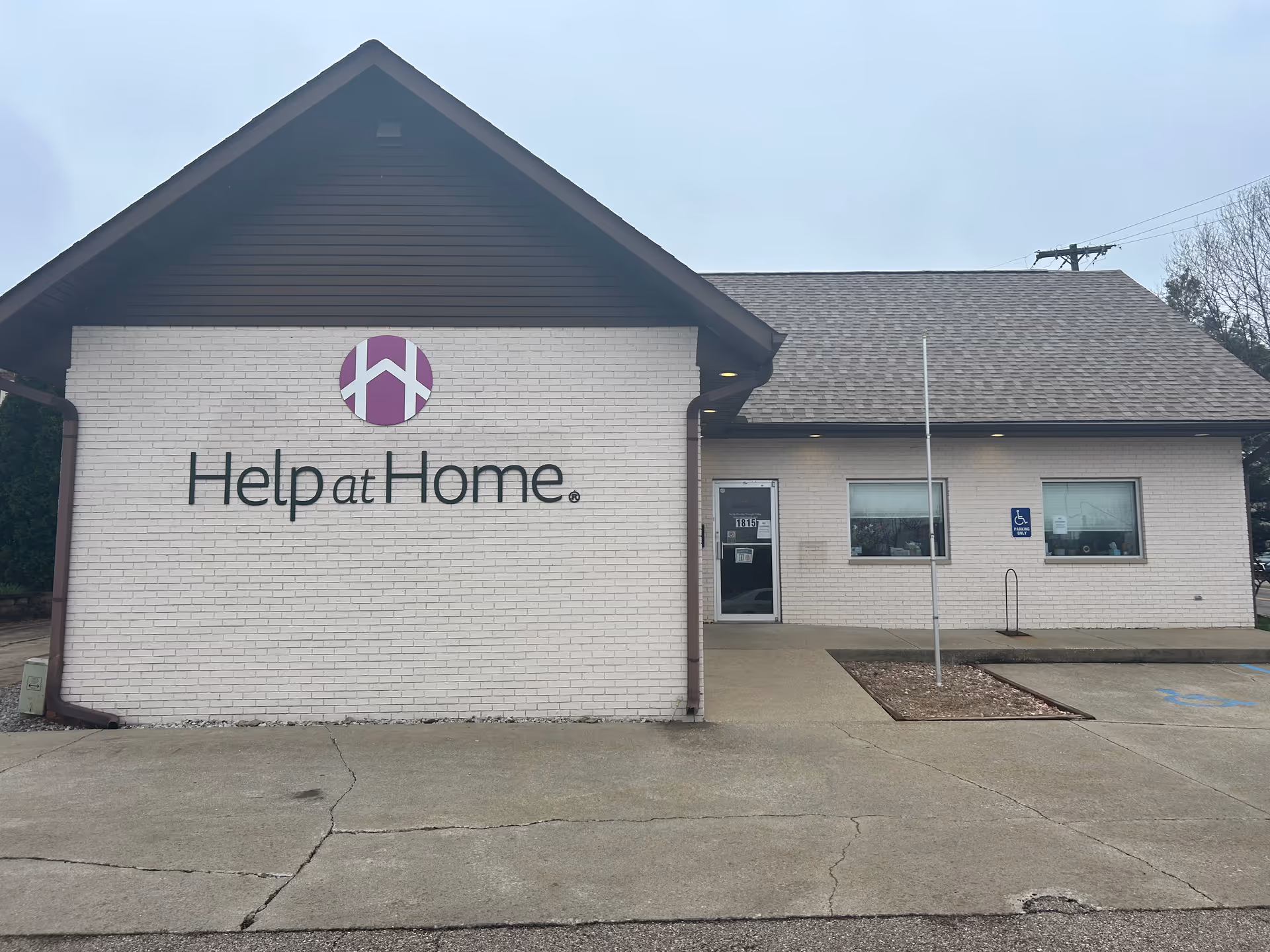 White brick facility with Help at Home sign, peaked roof, entrance door, and parking.