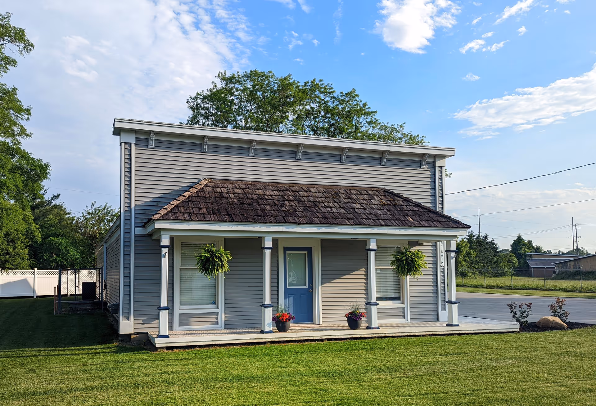 A light-gray two-story building with a front porch and hanging plants.