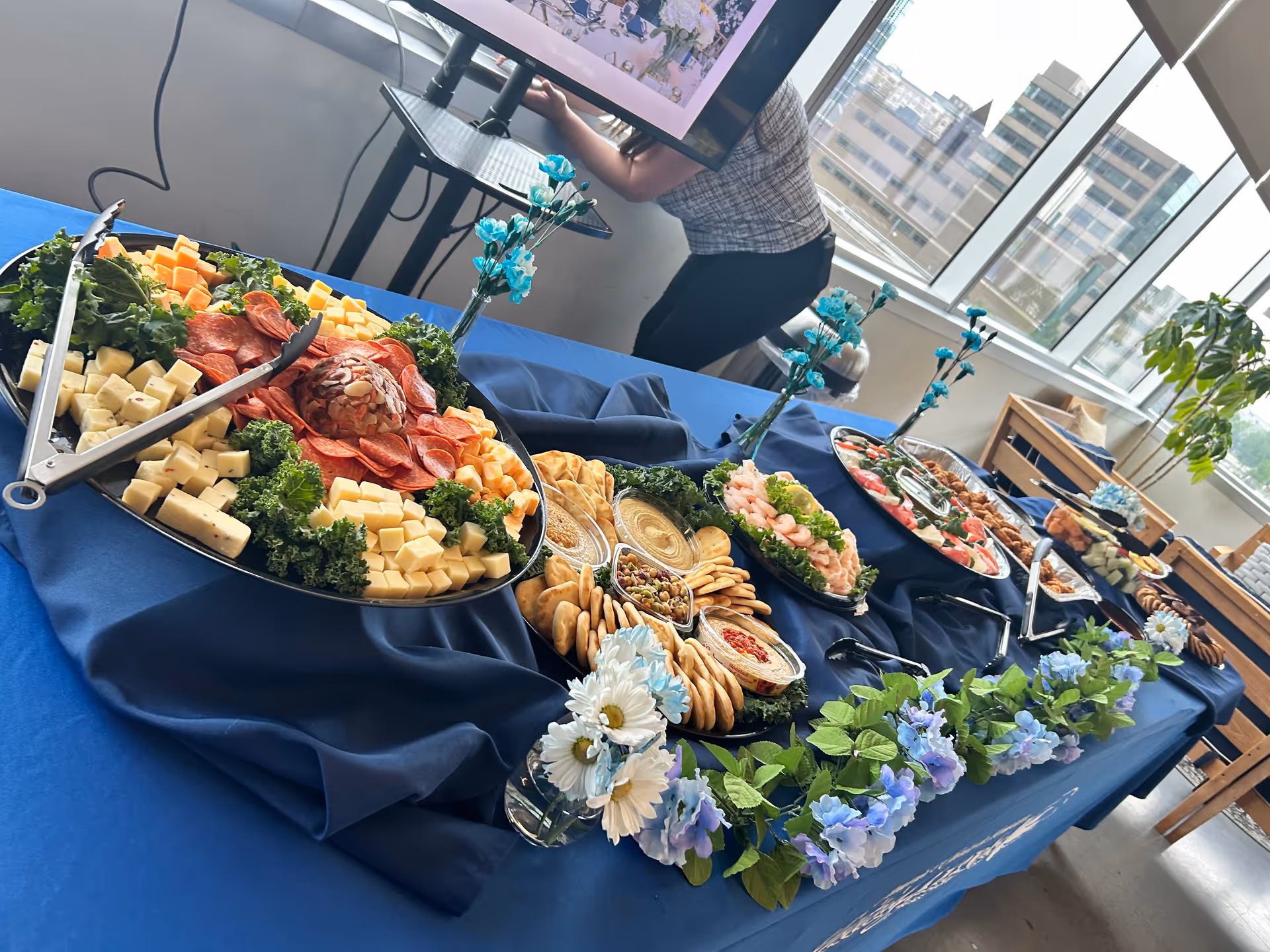 Buffet table with assorted cheeses, crackers, and toppings on a blue cloth in a dining area.