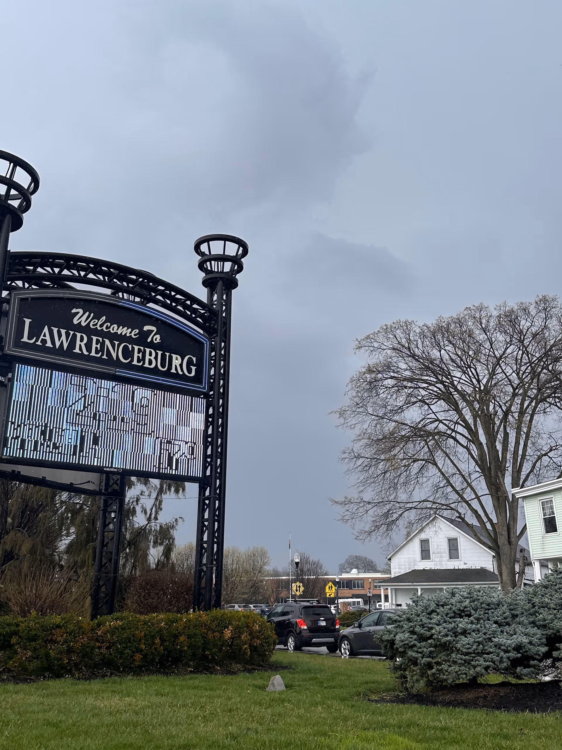 Large black welcome sign reading Lawrenceburg beside parked cars and trees under a cloudy sky.