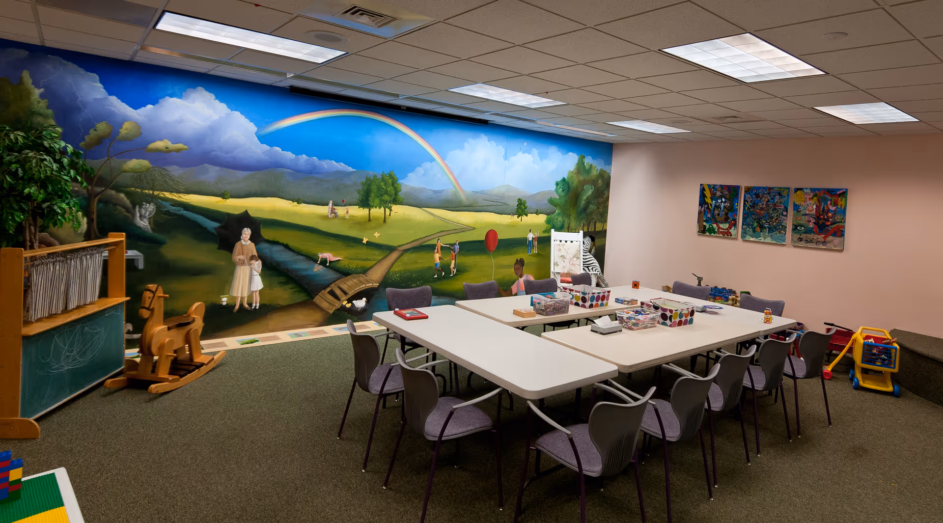 Colorful playroom interior with a large mural, tables, chairs, and toy supplies.