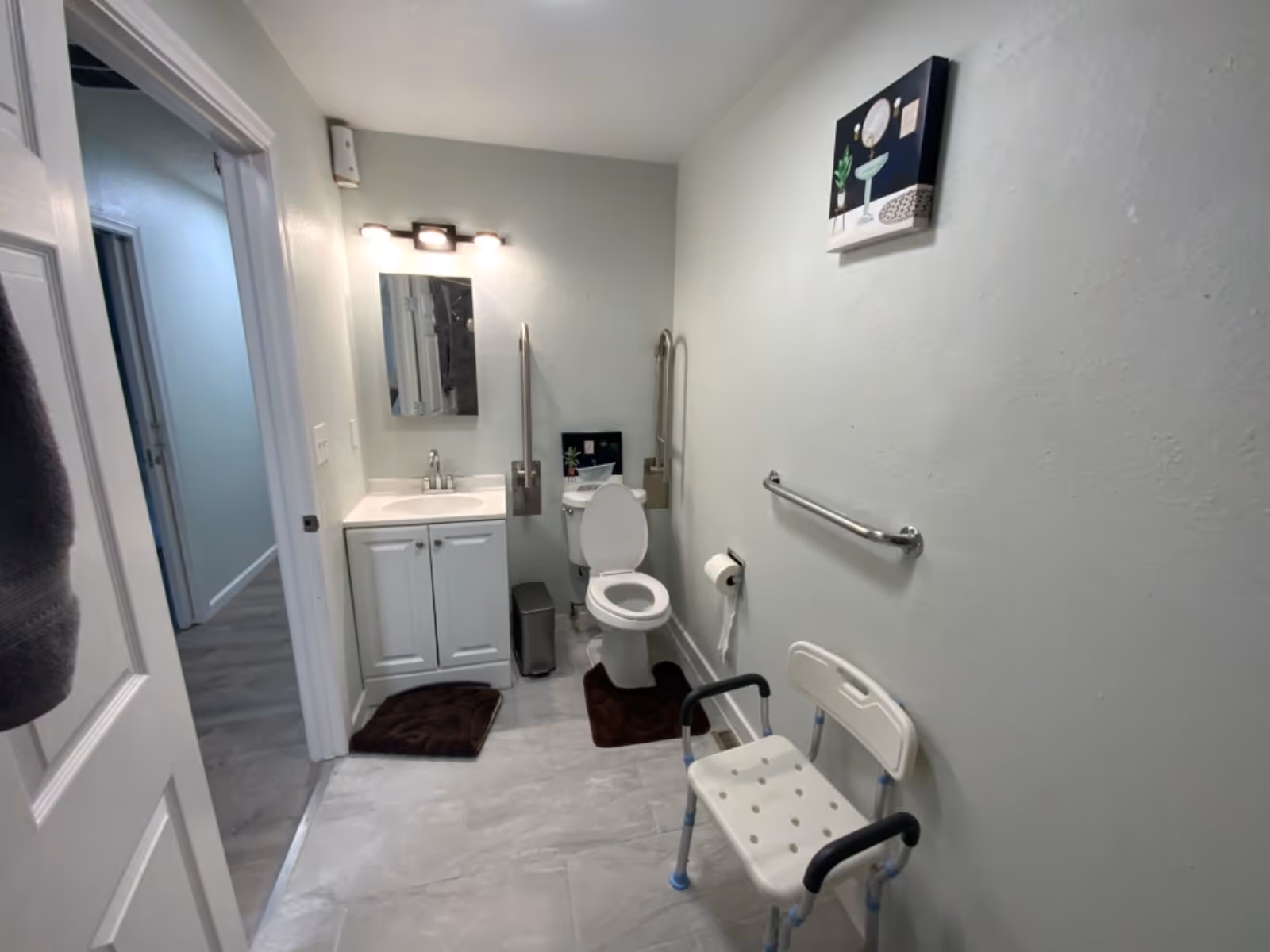 An accessible bathroom in a senior facility with sink, toilet, grab bars, and a chair.