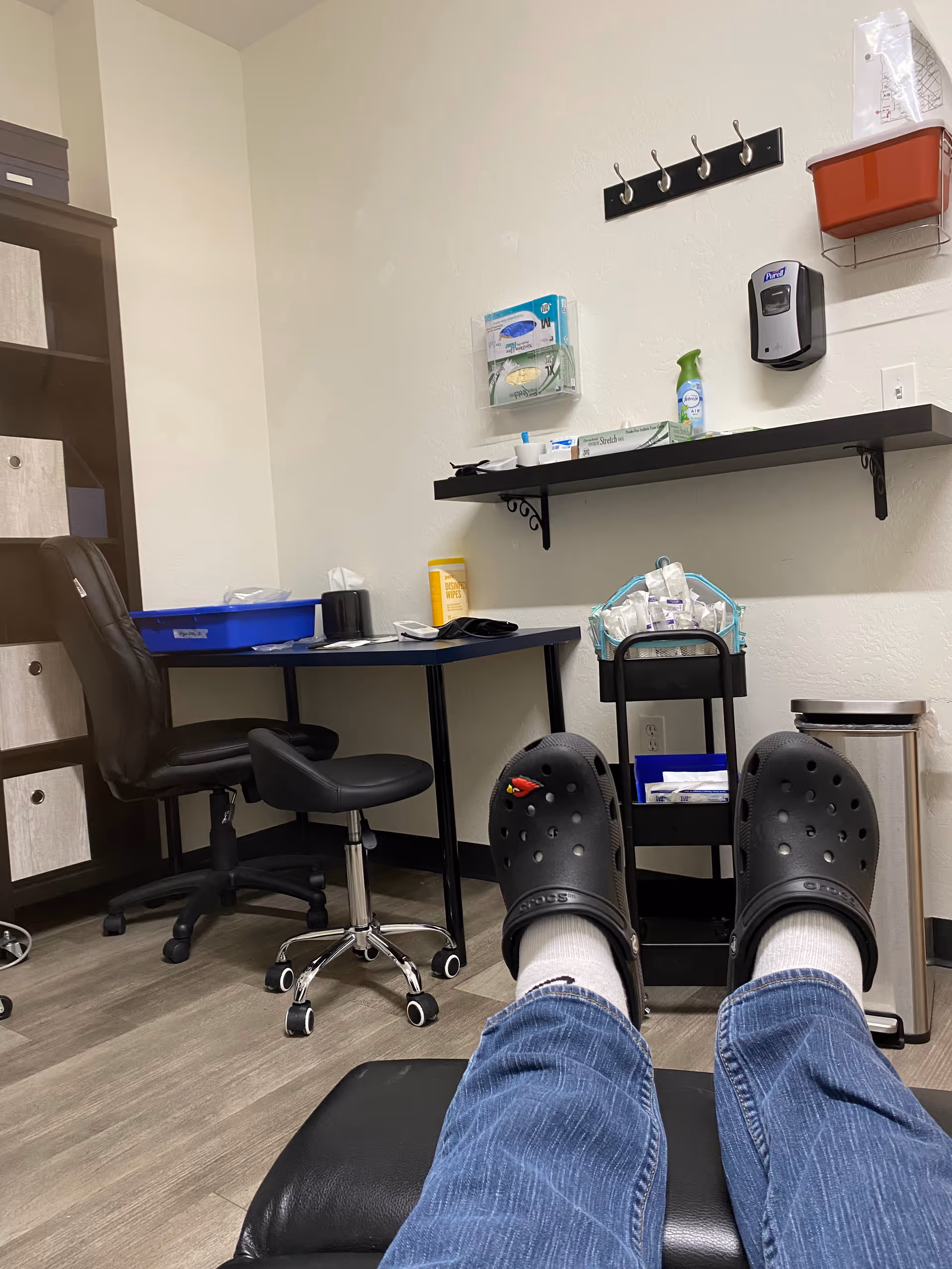 First-person view of a clinic room with feet in Crocs resting on a chair.