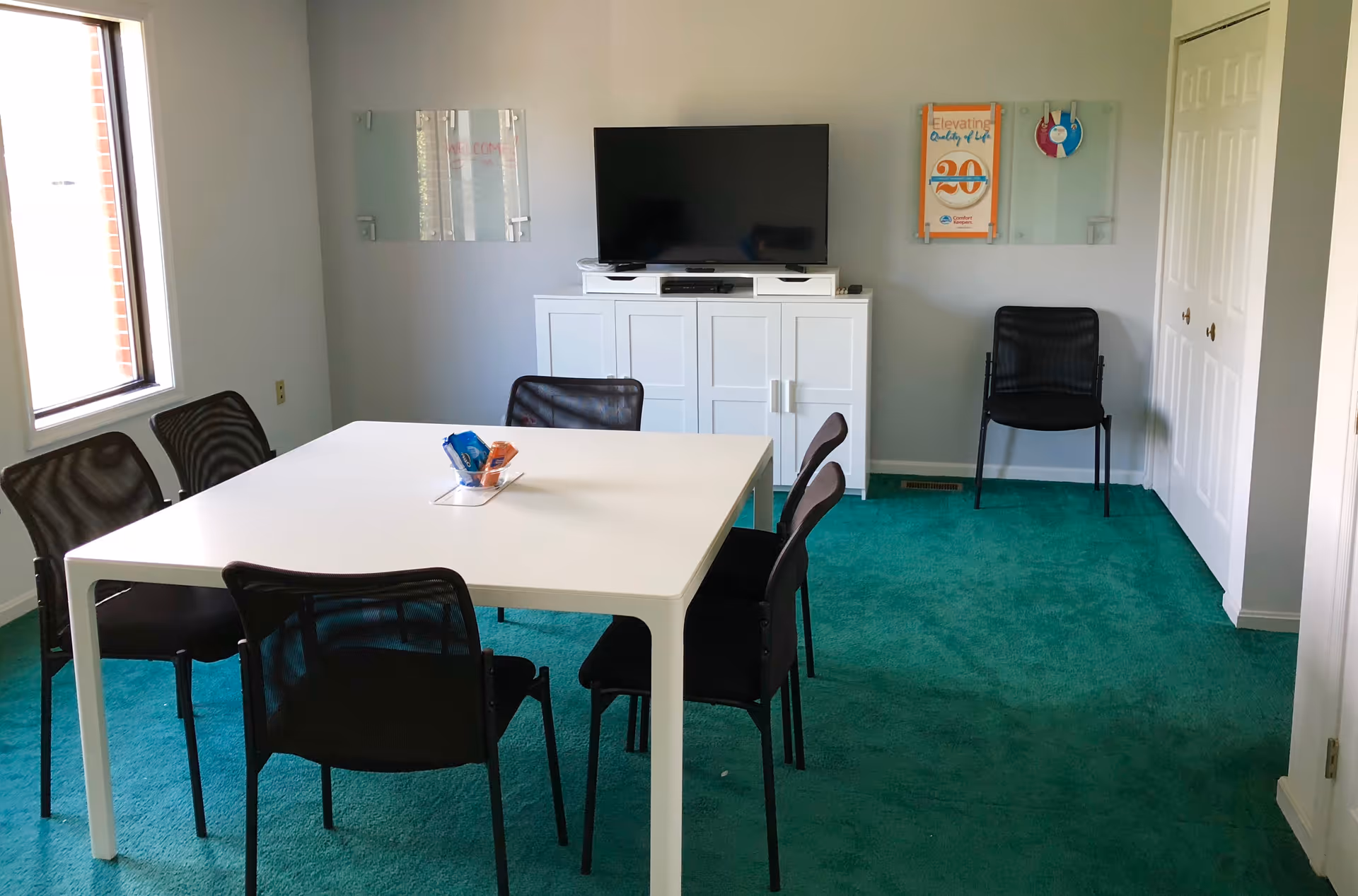 A bright room with a white rectangular table, black chairs, and a TV on a cabinet.