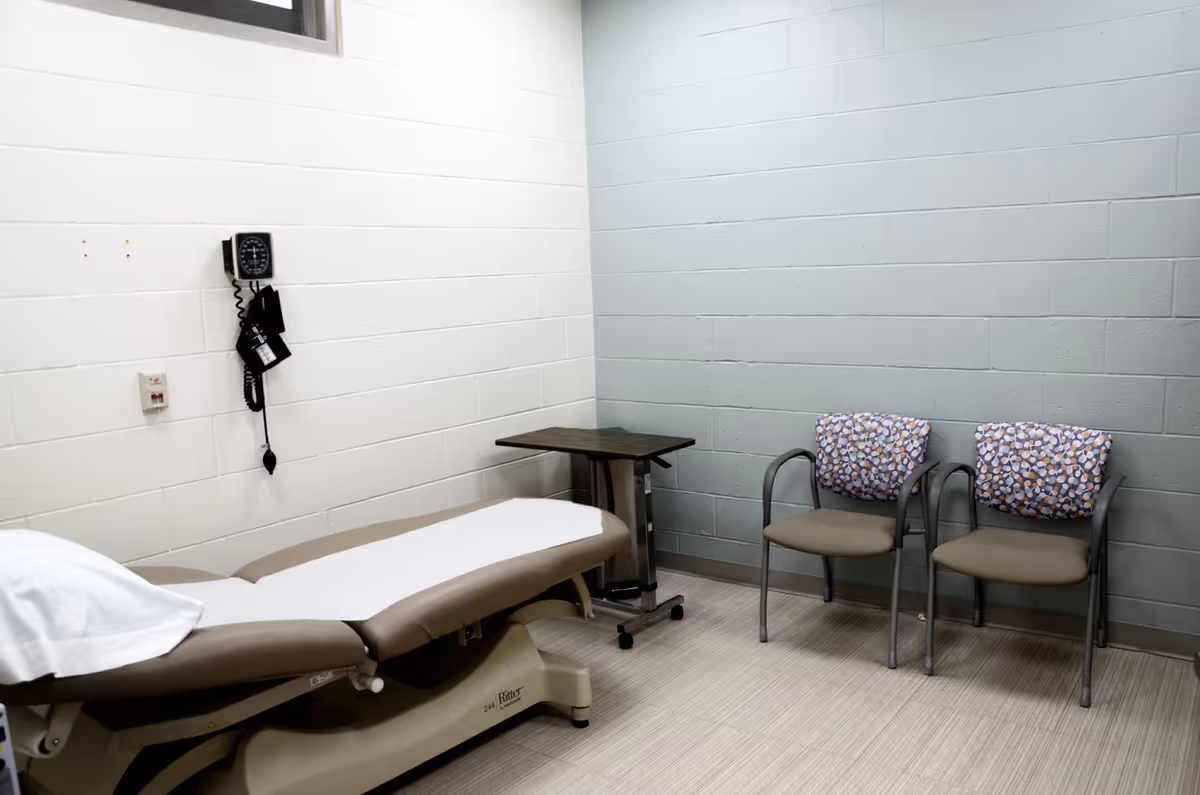 A medical examination bed with a small table and two patterned chairs in a clinical room.