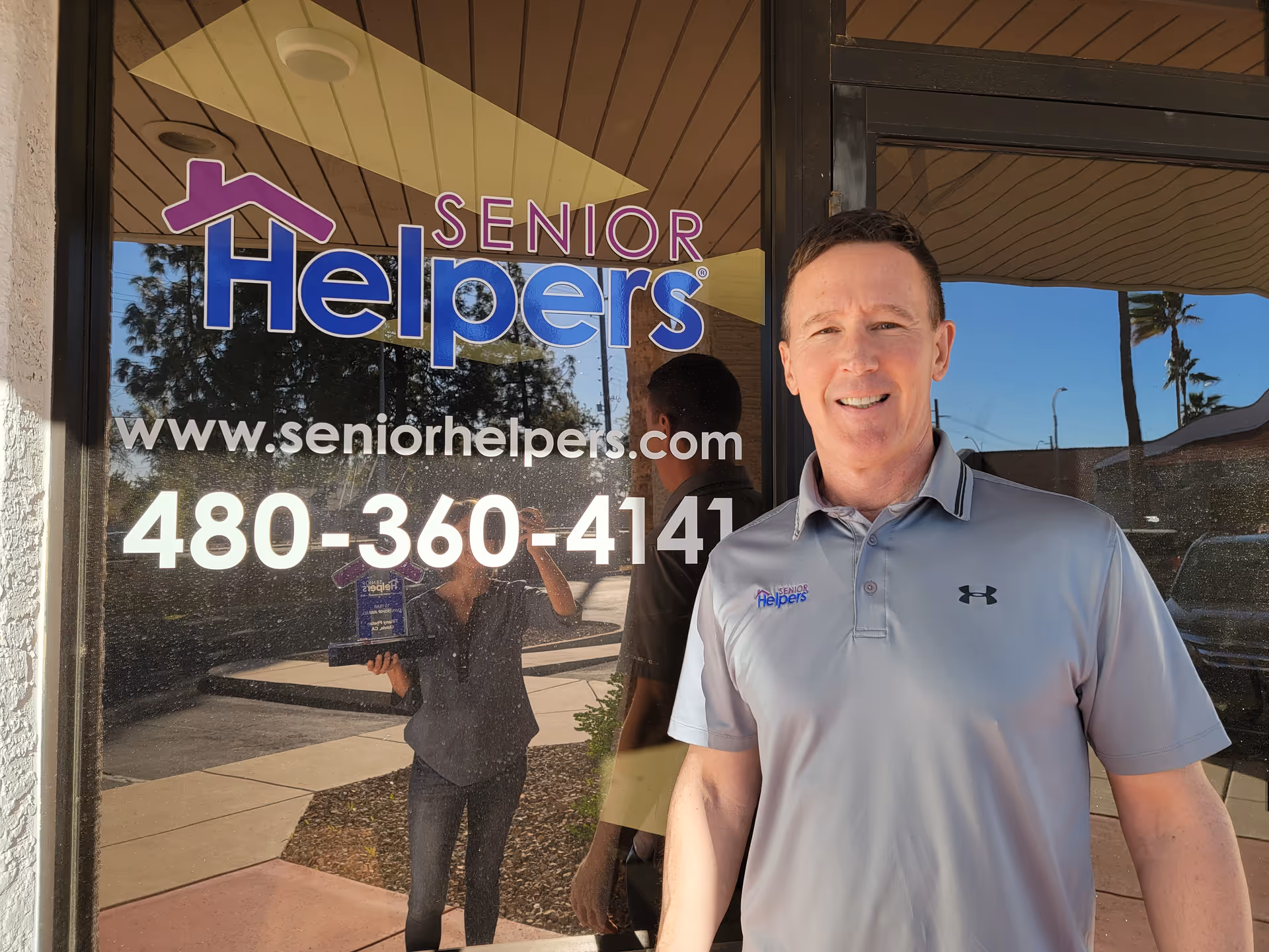 Man stands outside Senior Helpers storefront with logo and contact number on glass.