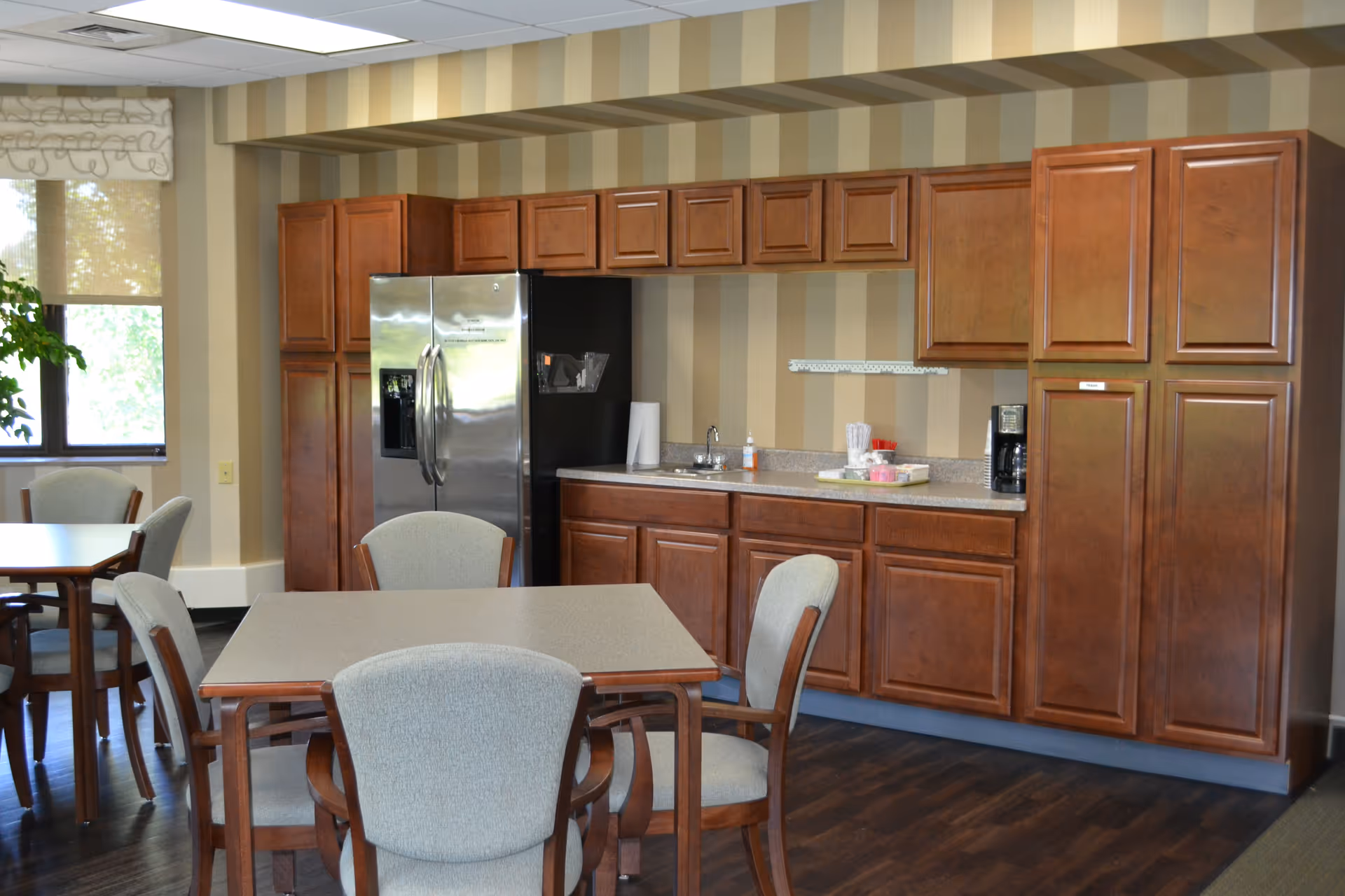 A communal kitchen area with wooden cabinets, a stainless fridge, sink, and dining tables.