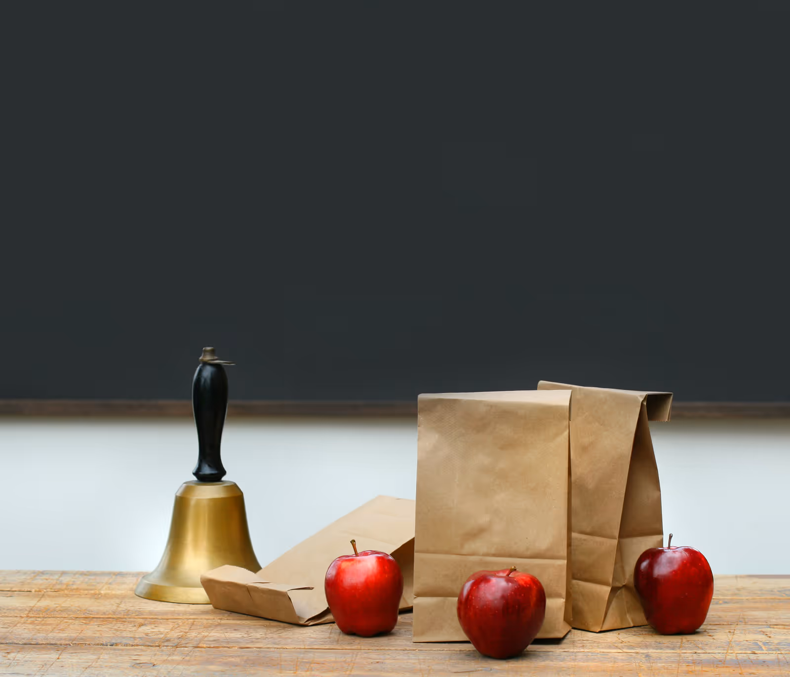 A wooden table with paper bags, red apples, and a bell against a dark backdrop.