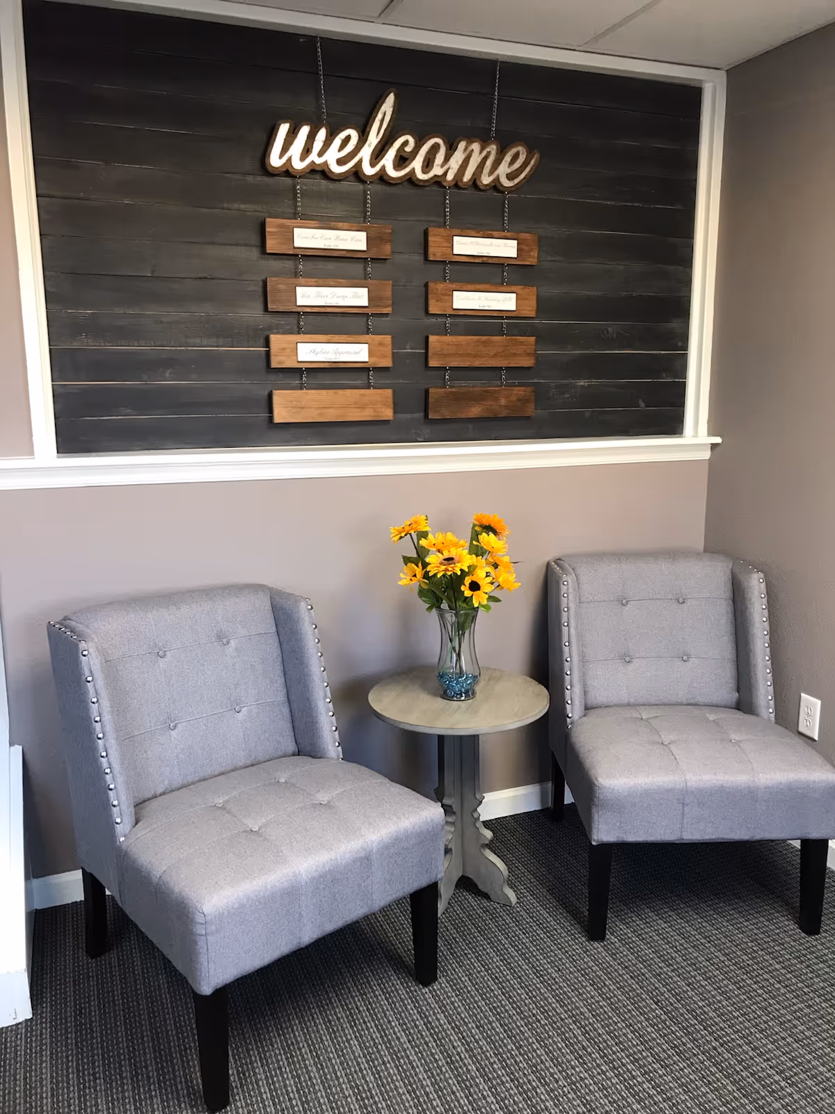 Two gray upholstered chairs flank a round table with a vase of sunflowers in a lobby.
