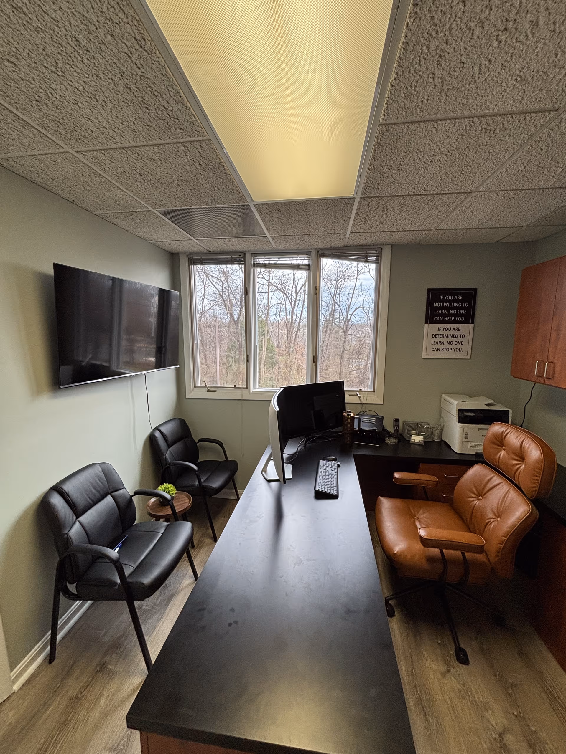 Interior office in a facility featuring a long desk, computer, chairs, printer, and a window view.