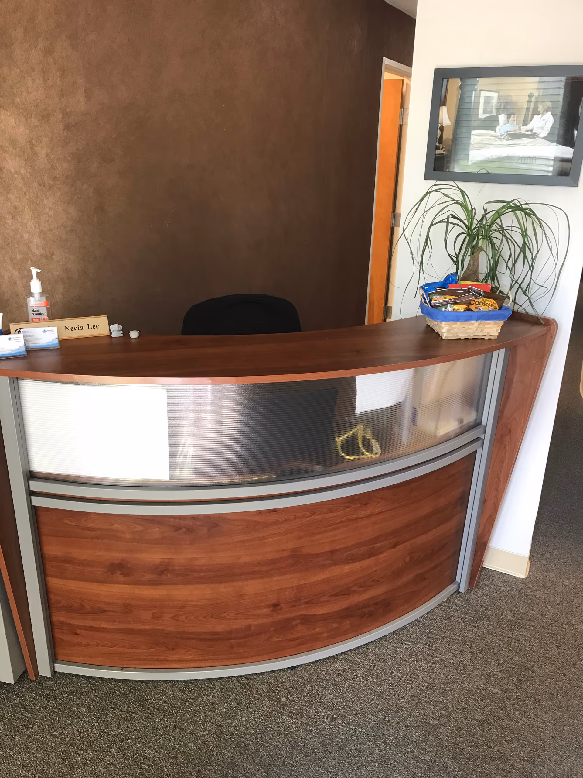 Curved wooden reception desk in a senior living facility lobby with a plant and snacks.