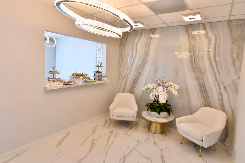 A modern marble-walled lounge with two white chairs, orchid, and dessert display near a serving window.