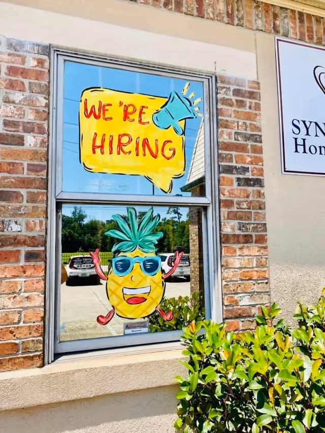 Front window of a brick building displays a bright We're Hiring sign and pineapple cartoon.