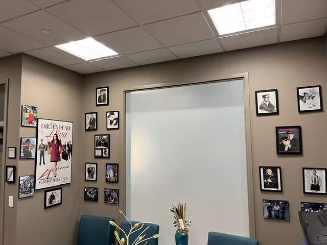 Beige lounge area with teal chairs, wall of framed photos, and frosted glass door.