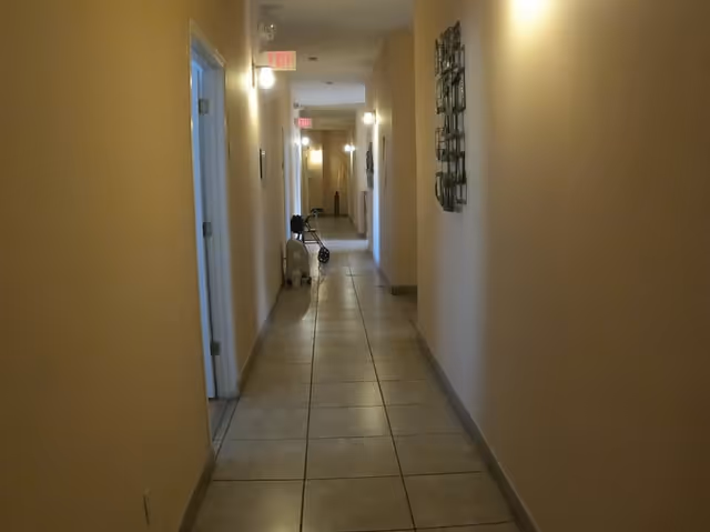 Long beige hallway in a senior facility with doors, tiled floor, and a cart.