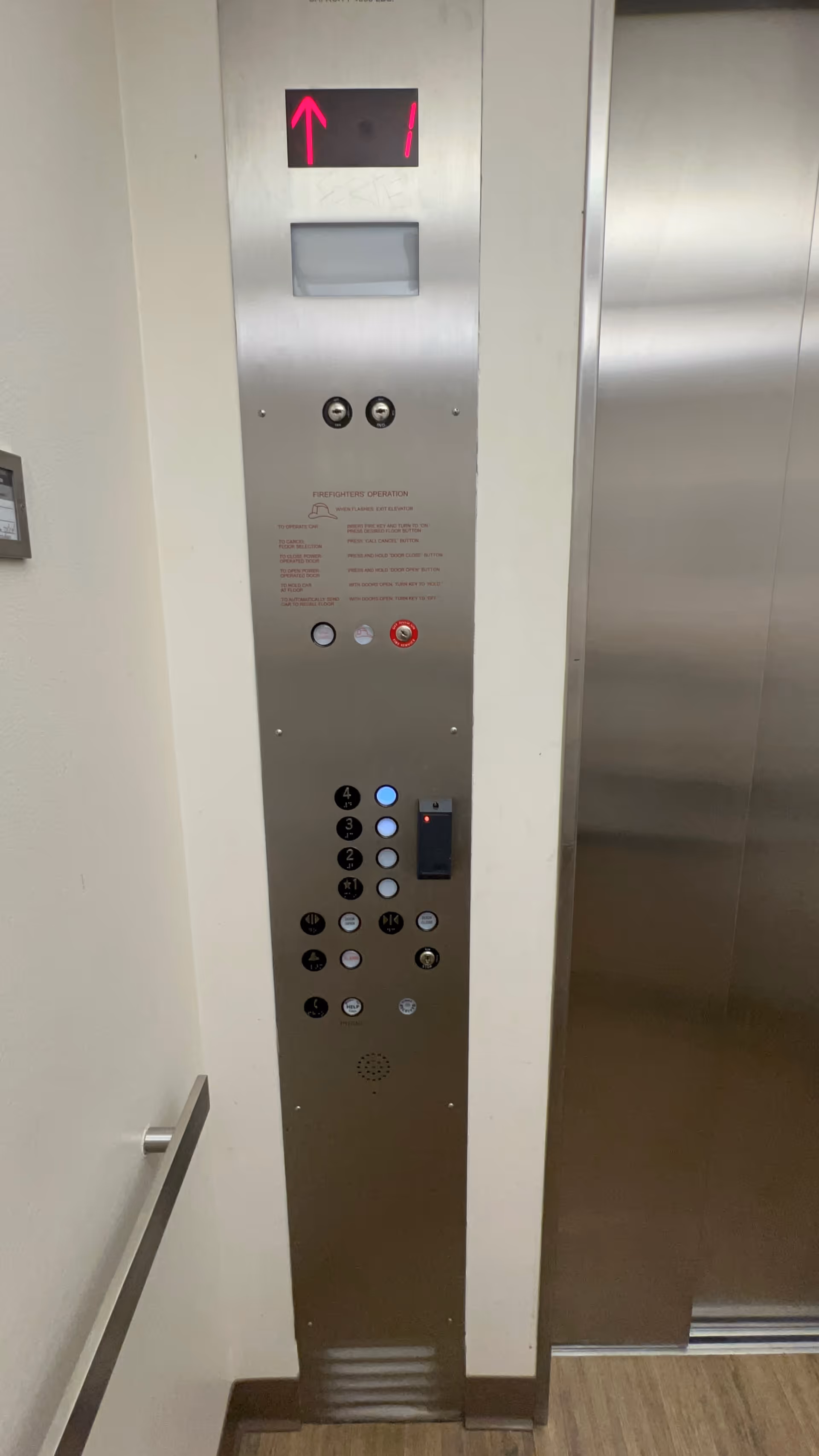 A stainless elevator control panel with floor buttons and a display in a hallway.