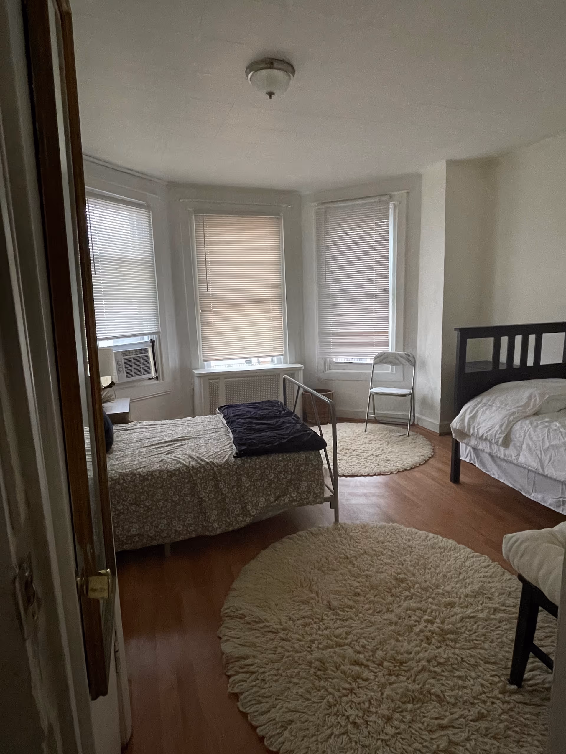 Sparsely furnished bedroom with two beds, blinds, chairs, and round rugs.