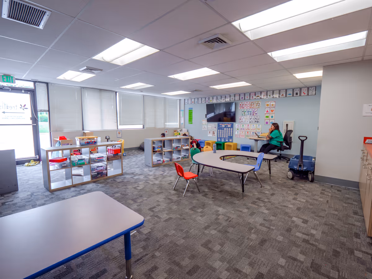 Bright indoor activity room with tables, chairs, shelves, and a staff member at a desk.