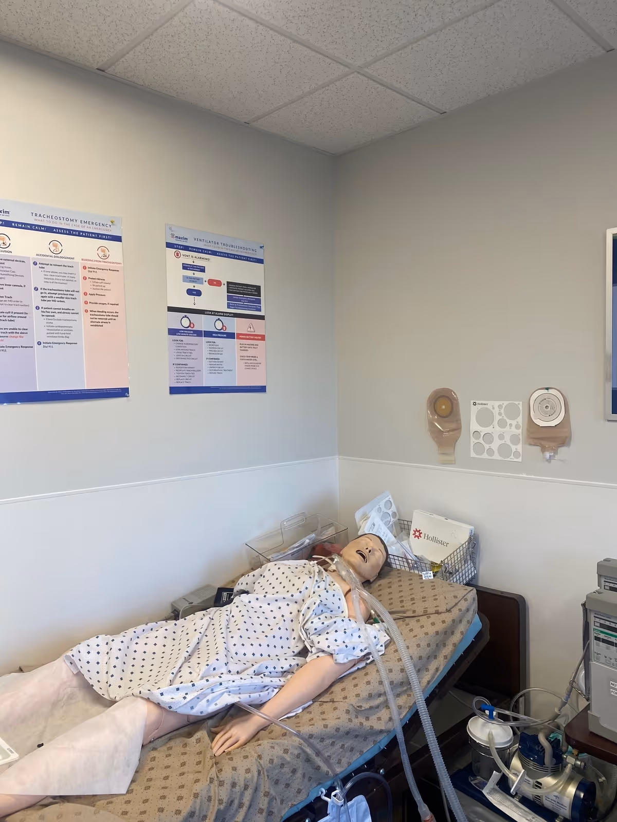 Medical training mannequin on a hospital bed in a clinical room with posters.