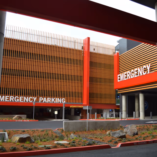 Exterior view of a hospital emergency department entrance with bright red signage and surrounding parking.