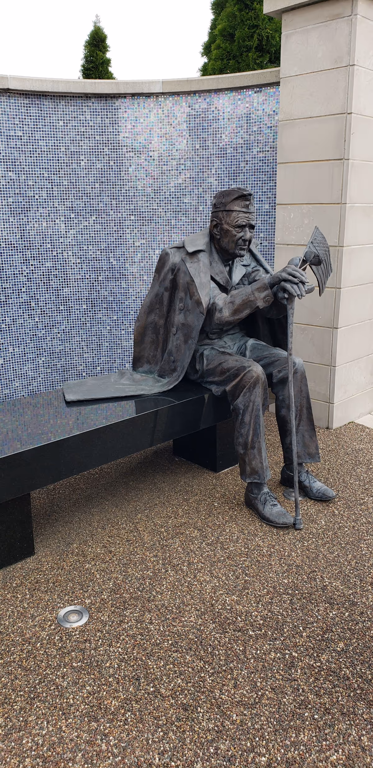 Bronze statue of a man with a cane sits beside a curved tiled wall outdoors