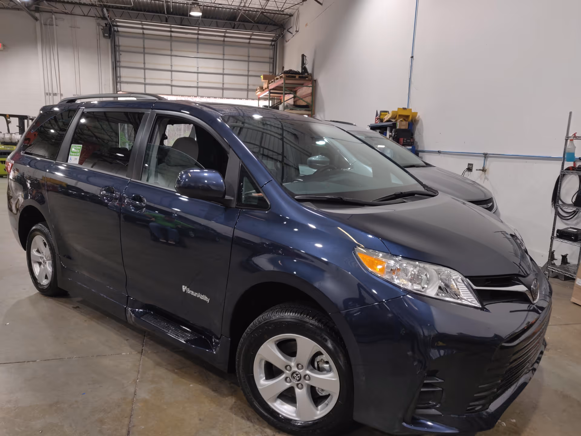 Blue mobility minivan inside a warehouse-style service area with shelves and equipment.