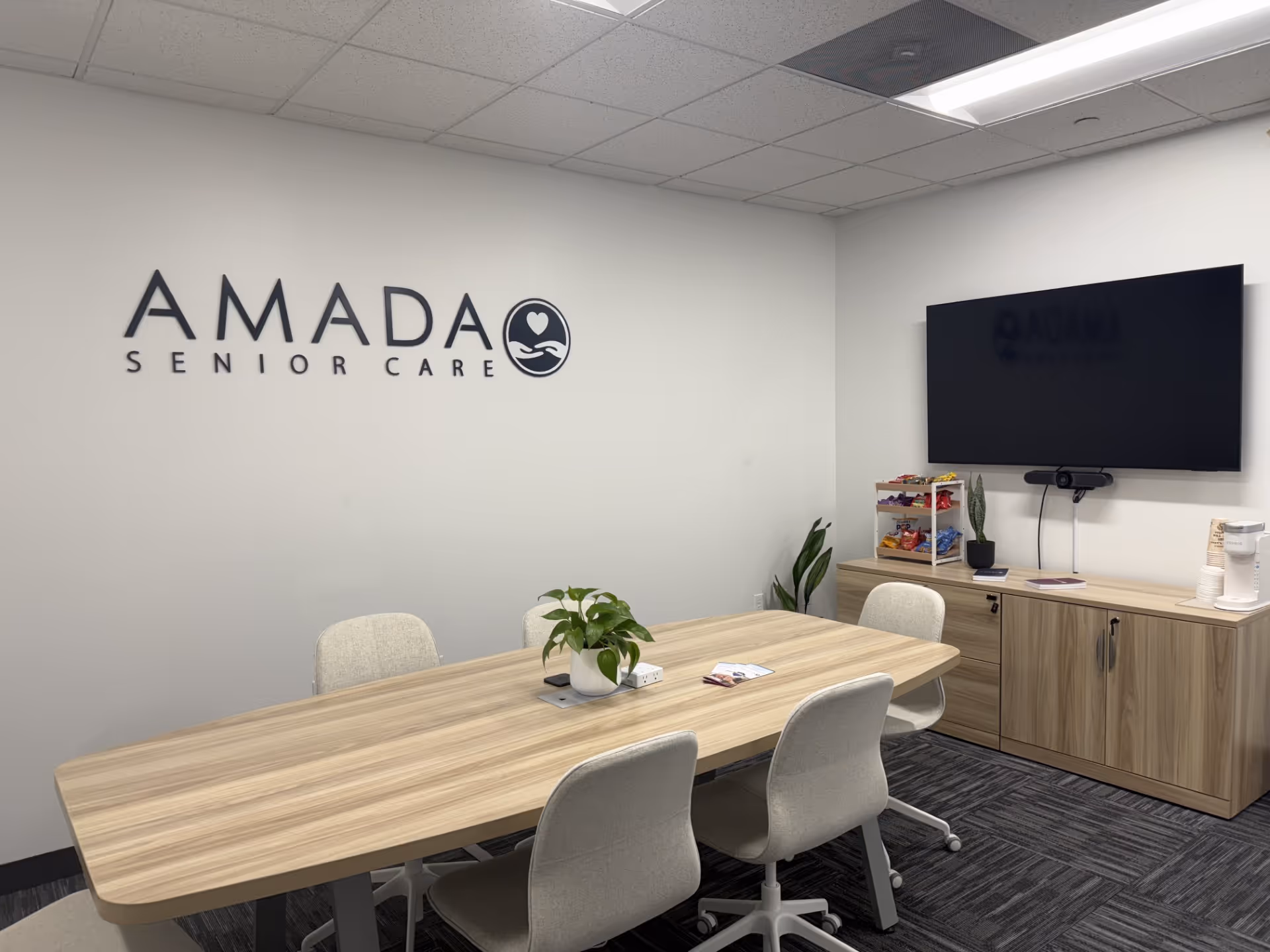 Amada Senior Care conference room featuring a long wooden table, chairs, TV, and snack station.
