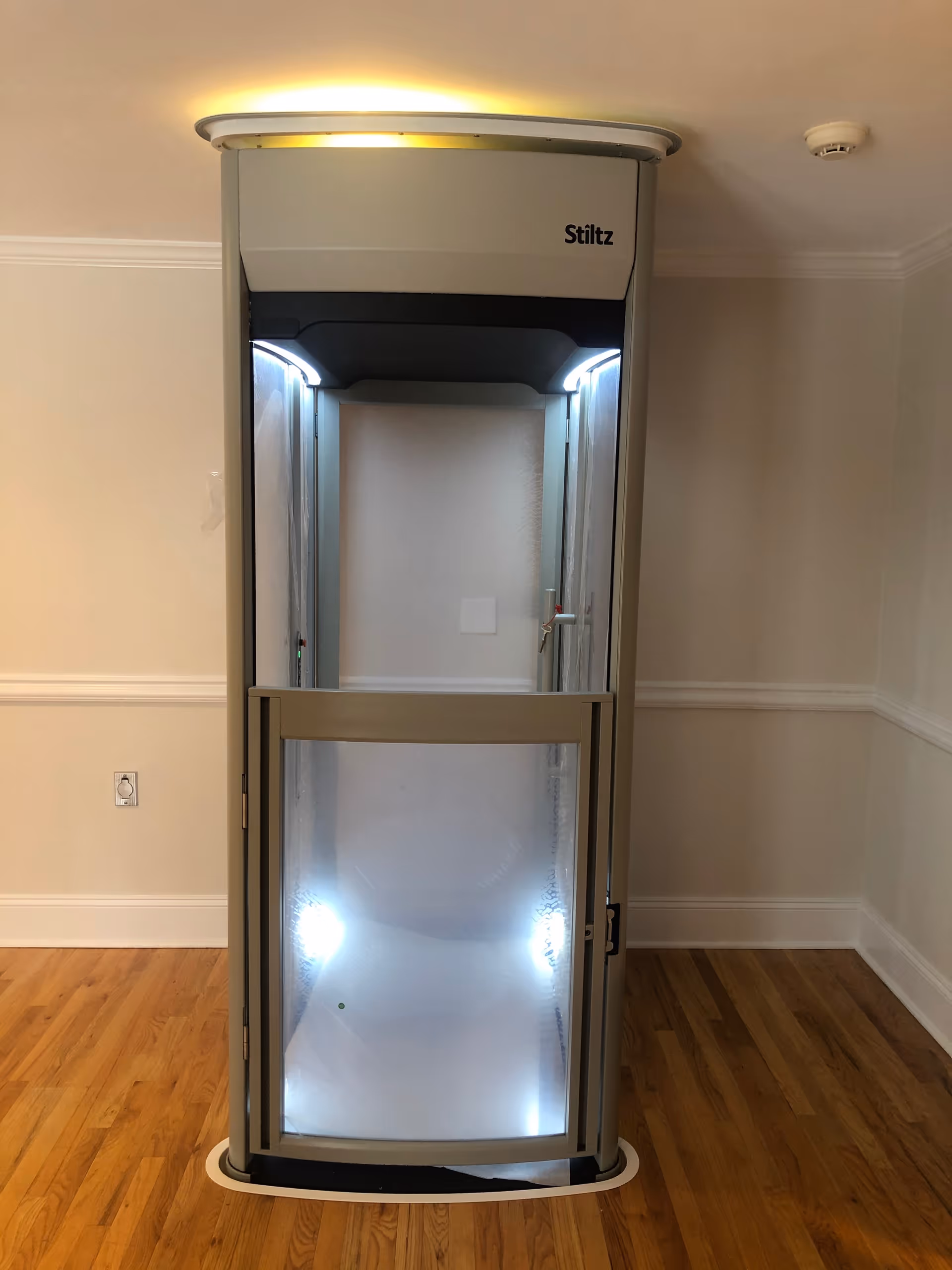 A Stiltz residential elevator sits in an interior hallway with wood flooring.